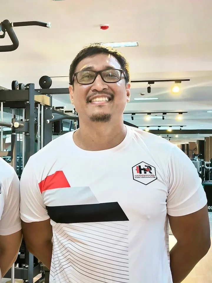 Coach Roger Justine Potot smiling in an H&R training shirt on the gym floor